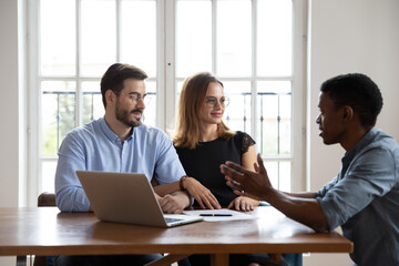 Male African American real estate agent talk consult young Caucasian couple show house dwelling plan on laptop, biracial man realtor speak have consultation with newlywed husband and wife in office