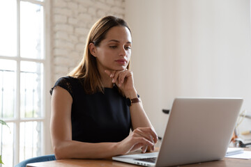Pensive Caucasian businesswoman sit at desk in office look at laptop screen thinking pondering, thoughtful young female employee busy working on computer watch webinar browse Internet