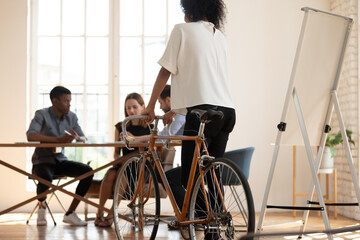 Young biracial female employee ride bicycle in modern office, back view of african American woman worker test try new bike, colleagues brainstorm discuss business ideas, stress free concept - Powered by Adobe