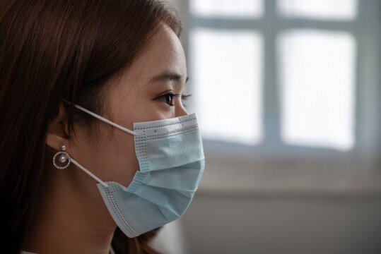 Woman in a medical mask at hospital and window background. Close-up of a young woman with a surgical mask on her face against coronavirus or COVID-19 disease. Medical and health protection concept.