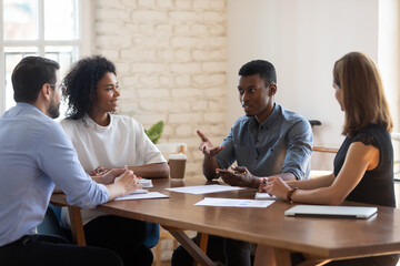 Multiethnic young colleagues sit at office desk brainstorm cooperate at briefing together, focused diverse multiracial businesspeople speak discuss ideas at meeting, collaboration, teamwork concept