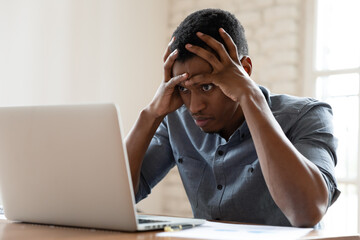 Mad African American young male employee look at laptop screen having operational software problems, angry biracial man worker work in office annoyed with slow Internet connection on computer - obrazy, fototapety, plakaty