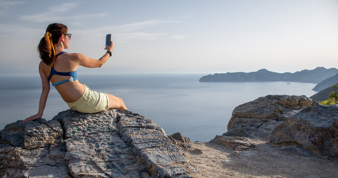A woman taking pictures with her phone of the sea view over a mountain