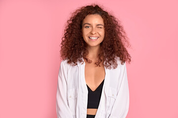 Photo of pretty cheerful girl with red curly hair smiling looking at camera over pink background.