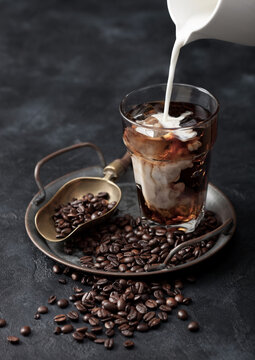 Pouring Milk Into Glass With Iced Black Coffee On Tray With Coffee Beans And Scoop On Black Background.