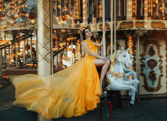 Fototapeta premium Young beautiful stylish woman sits astride a toy horse, rides a carousel. Long yellow bright dress fluttering in motion