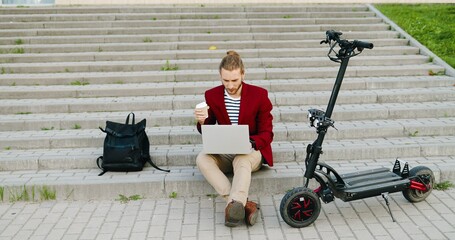 Young caucasian handsome man in red jacket sitting on steps outdoors, working on laptop computer,...