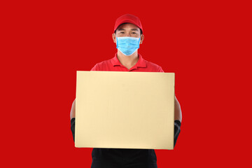 Happy young Asian delivery man in red uniform, medical face mask, protective gloves carry cardboard box in hands on red background. Delivery guy give parcel shipment. During COVID-19 outbreak