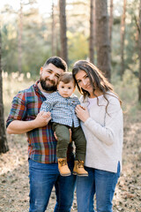 Happy family, dad, mom and little son, wearing stylish casual clothes, walking in the autumn pine forest at beautiful sunny day. Joyful parents holding little baby son and smiling to camera