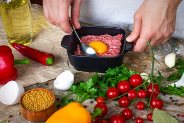 Egg yolk on minced pork in a black plate on a wooden table with cherry tomatoes, garlic, spices and mustard seeds. The process of cooking minced meat, cutlets