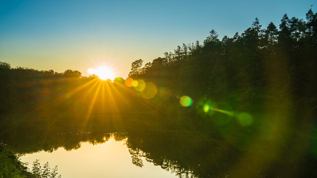 Germany, Stuttgart, Magical sunrays behind tree and forest silhouette reflecting in water of lake baerensee