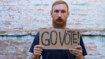 Naklejka na ściany i meble Man shows cardboard with Go Vote sign on brick wall urban background. Voting concept. Make the political choice, use your voice. Bearded guy calls to go to the presidential elections.