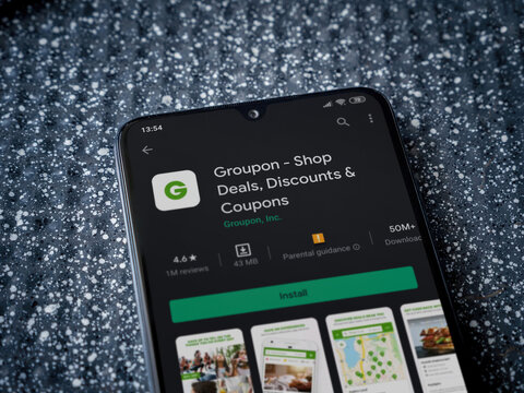 Lod, Israel - July 8, 2020: Groupon app play store page on the display of a black mobile smartphone on a metallic background. Close up top view flat lay.