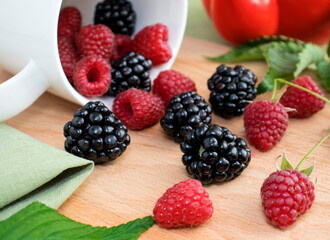 Raspberry and blackberry berries are spilled from a white cup on a wooden background. Nearby are green raspberry leaves and a teapot. The concept of making berry healthy tea. A healthy dessert.