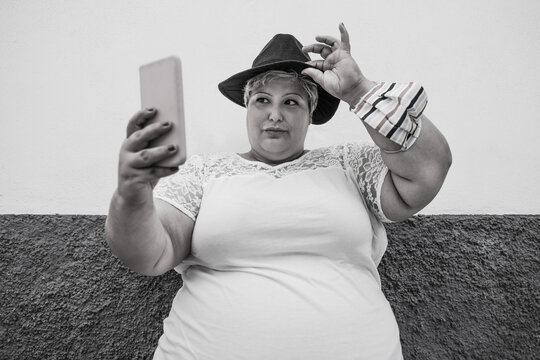 Plus size influencer girl taking a selfie with smartphone - Curvy woman with hat looking serious - Black and white image