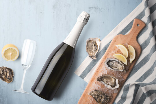 An unopened champagne bottle with a single glass on a blue tinted wooden background with fresh oysters and lemon on a wooden cutting board