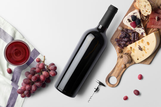 A picnic scene of red wine, cheese, nuts, meat, and fruit on a white background. A glass or wine rests on a towel with corkscrew and red grapes scattered around.