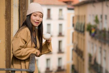 young beautiful and happy Asian Chinese woman in winter hat enjoying city view from hotel room balcony in Spain during holidays trip in Europe drinking coffee relaxed