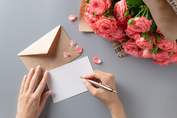 woman writing greeting card on gray table with bouquet of pink roses. Writing letter concept. top...
