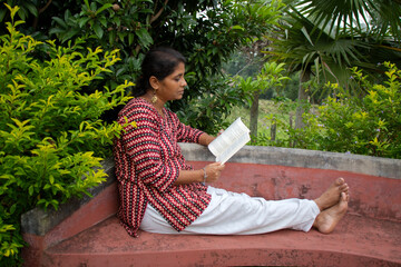A beautiful woman in casual clothing, sitting on a red bench is reading a book and thinking about the story in a park 