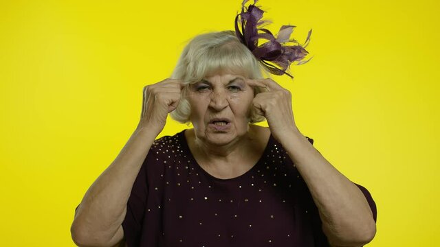 Displeased annoyed senior old woman showing stupid gesture. Elderly grandma on yellow background