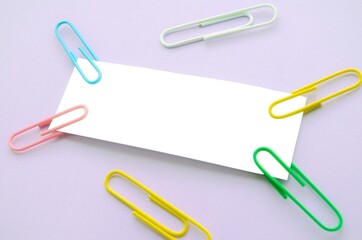 white paper sheet and colored paper clips on purple background. Copy space for text. Stationery things. Colored paper clips hold paper. Space for writing notes.