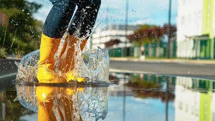 LOW ANGLE: Girl in bright yellow rubber boots jumps into the glassy puddle.