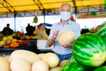 Fototapeta premium retired european man wearing medical mask protecting against the virus selling melons and watermelons in market
