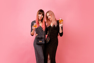 Stylish girls with glasses of alcohol drinks posing on light pink background. Party mood. Wearing luxury sequins dress .