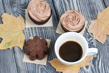 Chocolate cupcakes with cream cheese cream. Nearby is a cup of coffee and dried maple leaves. On pine planks painted black and white.