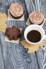Chocolate cupcakes with cream cheese cream. Nearby is a cup of coffee and dried maple leaves. On pine planks painted black and white.