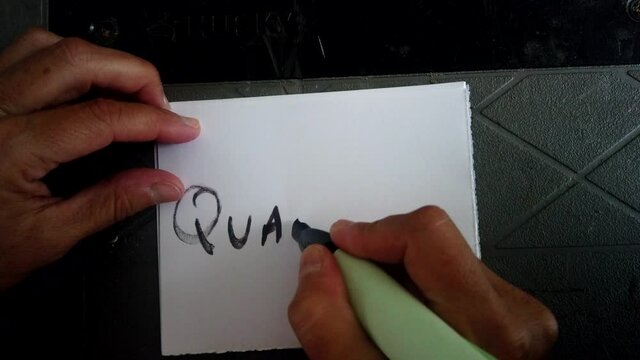 Adult Caucasian female hand writes with black magic marker and scribes in block capital letters word quarantine on white piece of square paper, covid-19 shutdown and pandemic, directly above static
