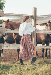 Beautiful slender girl of model appearance dressed in a shirt, skirt and boots posing in the stable near the horses