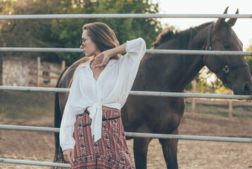 
a woman of model appearance with long hair is dressed in a shirt, a long skirt stands near the fence on the background of an equestrian farm near the horses at sunset