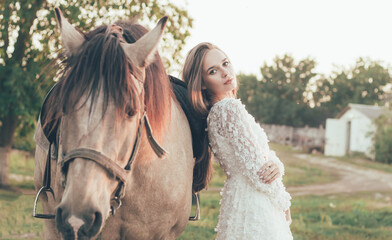 
A woman of model appearance in a white dress stands near a horse in a field