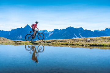 Naklejka na ściany i meble nice woman riding her electric mountain bike the Three Peaks Dolomites, reflecting herself in the blue water of a cold mountain lake
