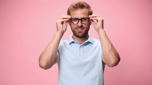 casual young man in blue polo shirt smiling, putting glasses on, nodding and crossing arms on pink background