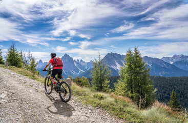 Plakat nice and active senior woman riding her electric mountain bike on a old military road from Toblach upt to the summit of Marchkinkele eith spectacular view to the Three peaks of Lavaredo, South Tirol, 