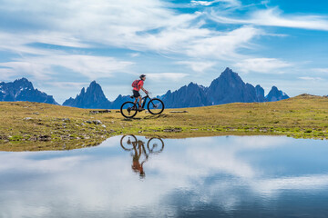 Naklejka na ściany i meble nice woman riding her electric mountain bike the Three Peaks Dolomites, reflecting herself in the blue water of a cold mountain lake