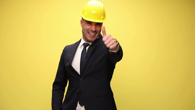 young businessman wearing a helmet, having one hand in pocket and making a ok sign with the other, smiling wide on yellow background