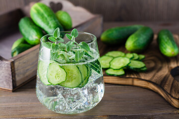 Detox drink. Water with pieces of cucumber, ice and mint leaves in a transparent glass and a cutting board with a vegetable on a wooden table