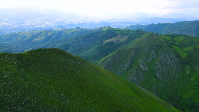 Grassy covered peak of a ridge in Asturias, Spain coming into shot as drone descends behind it showing carved crevasses in the rock down the other mountains, a blue haze filling the horizon.