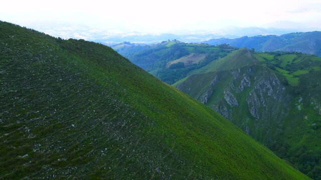 Crest of a mountainous ridge above Asturias, Spain revealing a steep valley running between the peaks, covered by coniferous forest and a mountain range filling the horizon beyond a blue haze.