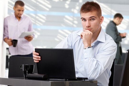 Serious young casual business team leader with laptop at office, sitting at table, colleagues in background, looking at camera, hand under chin.