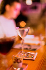 A lifestyle atmospheric photo of a cocktail in a nick and nora glass at the bar in the evening, people in the background, high iso, selective focus