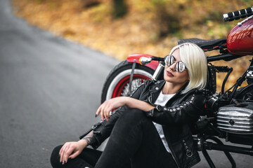 Fototapeta na wymiar Pretty blonde biker girl in sunglasses sitting near red motorcycle on the road