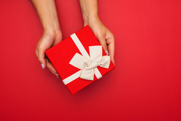 Female hands are holding present box or gift box with gold ribbon over red background. Top view