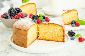 Homemade classic vanilla sponge cake or biscuit sprinkled with powdered sugar and fresh berries on top on a white plate on a light wooden background.