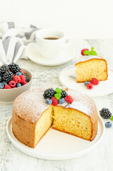 Homemade classic vanilla sponge cake or biscuit sprinkled with powdered sugar and fresh berries on top on a white plate on a light wooden background.