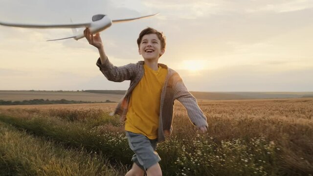 Happy boy holding airplane in his hand, runs across field of golden wheat against of sunset of summer day, throwing towards bright sun slow motion. Child plays lifestyle. Travel. Childhood